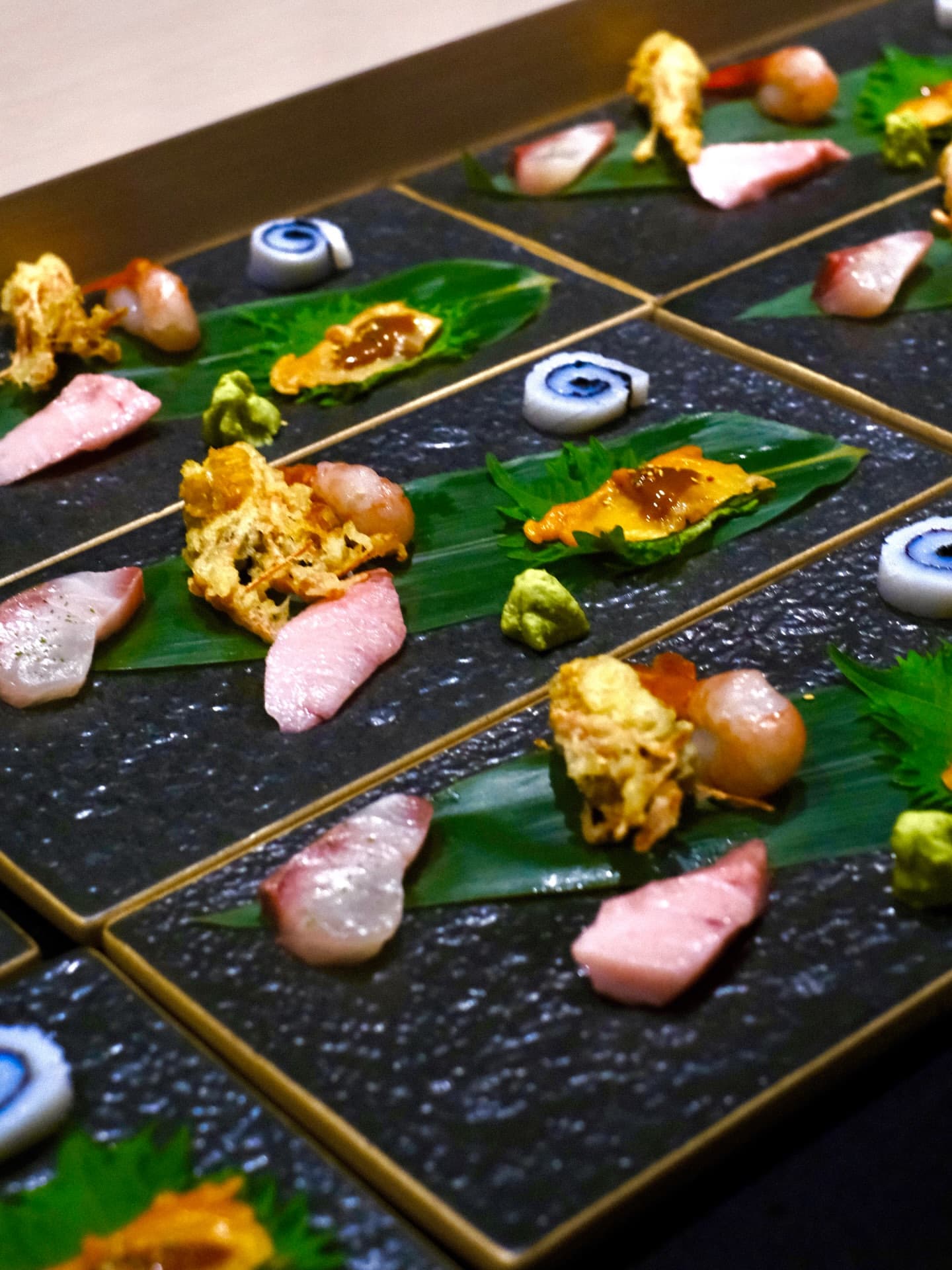 Rows of sashimi and small appetisers on black slate plates with wasabi and leaves