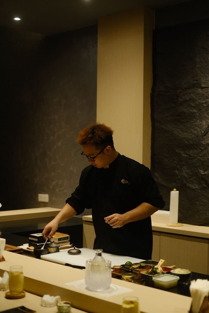 Alex, founder and itamae of DEN Culinary, in a black chef’s jacket and glasses, working at the counter with a knife, a stack of boxes and a row of garnish bowls in front of him