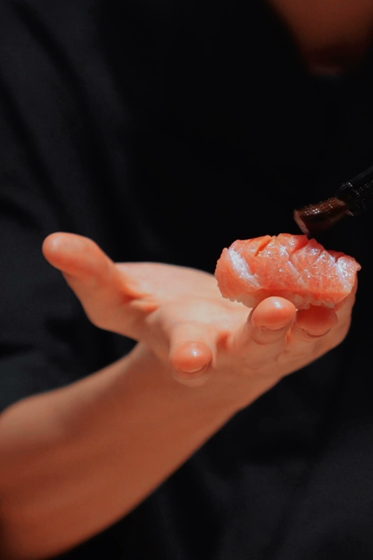 A chef’s open palm holding a piece of tuna nigiri while brushing it with soy