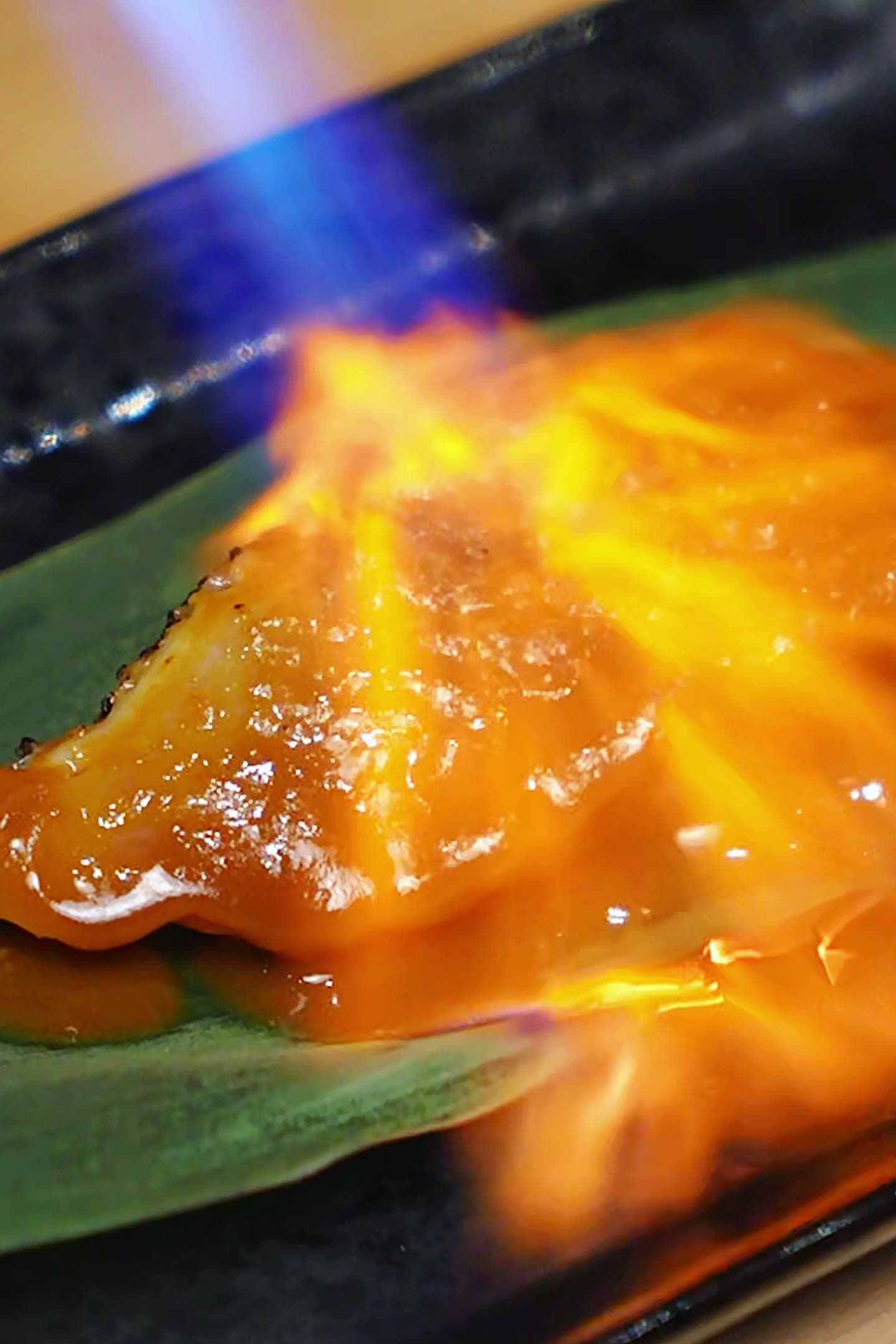A blowtorch flame searing a glazed fish fillet on a bamboo leaf