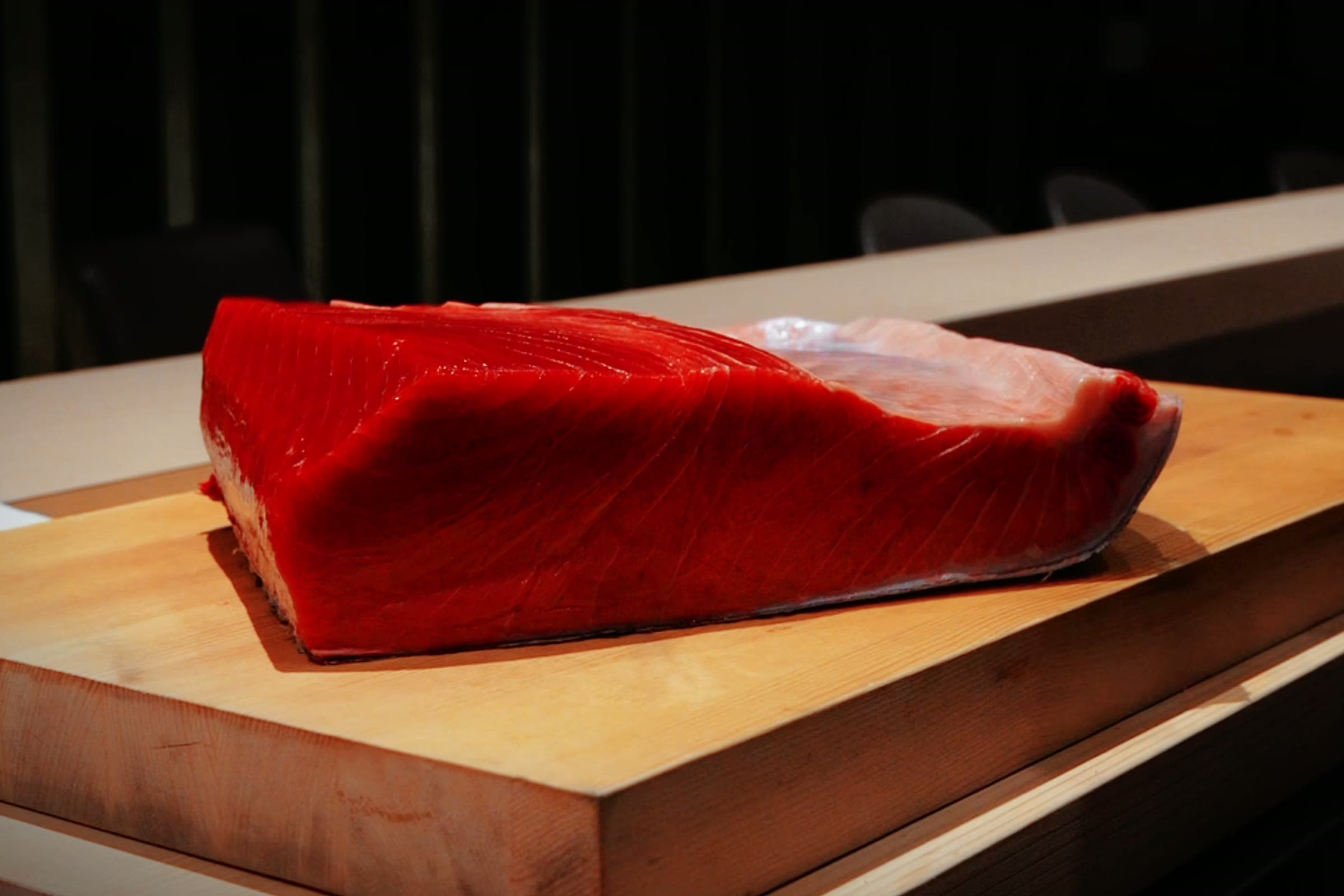 A whole loin of raw bluefin tuna on a pale wooden board at the counter, the dark dining room behind